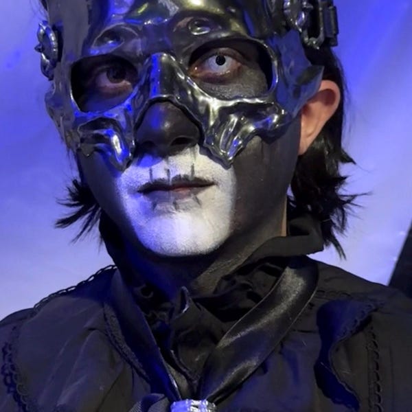 May include: A metallic silver resin Crypta mask with a skull-like design, featuring intricate molded details and side attachments. The mask is worn over a face painted with black and white makeup, paired with a black ruffled collar and a silver ribcage-style neck accessory.