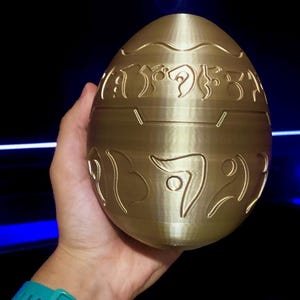 May include: A gold-colored egg-shaped object with intricate carvings and symbols. The egg is smooth and appears to be made of a hard material.