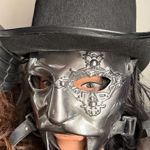 Op de afbeelding: Een metallic zilveren halfmasker met ingewikkelde reliëfdetails en filigraan rond het linkeroog. Het masker is bevestigd met zwarte lederachtige riemen en gespen en wordt gedragen onder een hoge, zwarte vilten hoge hoed met een brede rand.