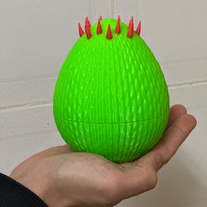 May include: A green egg-shaped object with a textured surface and red spikes on top. The object is being held in a person's hand.
