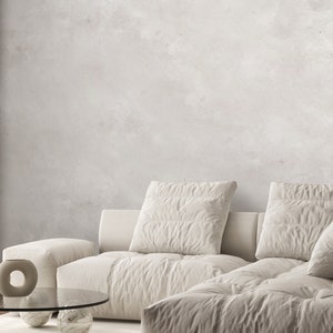 May include: A modern living room featuring a large, cream-colored sectional sofa with multiple pillows. A round glass coffee table with marble-like sphere legs sits on a light rug. The background is a textured, light gray wall.