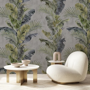 Puede incluir: Una habitación con un papel pintado de temática tropical con grandes hojas de plátano verdes y grises y frondas de palmera. Un sillón blanco y redondeado y una pequeña mesa auxiliar redonda con un jarrón decorativo están frente al papel pintado. Una mesa de centro blanca con patas doradas está en primer plano.