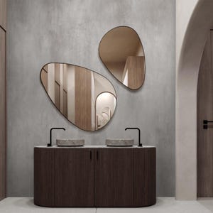 May include: Two irregularly shaped mirrors with black frames hang above a dark wood vanity with two stone sinks and black faucets. The vanity has a ribbed texture. The mirrors reflect the interior of a room with wooden panels.