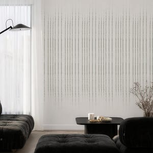 May include: A modern living room featuring a white wall with vertical gray brushstroke pattern wallpaper. The room includes a black sofa, ottoman, and coffee table. A black floor lamp and sheer white curtains are also visible.