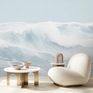 May include: A modern living room scene featuring a large wave mural in shades of blue and white. A white, curved armchair and two small, round tables with gold legs are in the foreground. Two beige bowls and a decorative vase are on the tables.