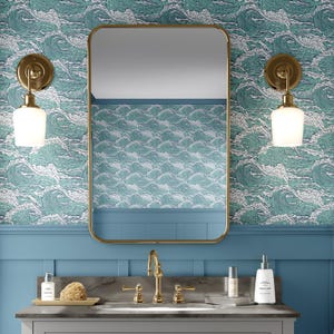May include: A bathroom scene featuring a rectangular gold-framed mirror, two sconces with white glass shades, and a sink with gold fixtures. The wallpaper has a teal and white wave pattern, and the walls are painted blue.