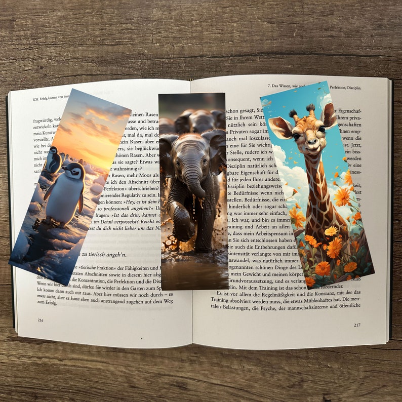 Drei Lesezeichen mit Tieren. je mit einem Pinguin, Elefant und einer Giraffe, in einem offenen Buch, liegend auf einem Holztisch.
