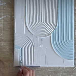 May include: A close-up of a white canvas with blue paint being applied to create a textured, abstract design. The design features a series of curved lines and shapes.