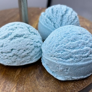 Op de afbeelding: Drie bollen lichtblauwe badbruisballen, die op ijs lijken, zijn gerangschikt op een houten oppervlak. De badbruisballen hebben een licht korrelig uiterlijk en zijn zacht pastelblauw.