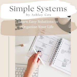 Op de afbeelding: Een wit bureau met een laptop, een toetsenbord en een hand die in een planner schrijft. De planner heeft de tekst "Simple Systems by Ashley Cox" en "Learn Easy Solutions to Organize Your Life". De planner heeft ook de tekst "Tidytribe.com" onderaan.