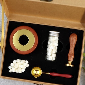 Puede incluir: Un kit de sellado de cera en una caja marrón. El kit incluye un sello de cera de madera y dorado, un mango de madera con un sello dorado, una cuchara dorada, un tarro de cuentas de cera blancas y un horno para derretir cera.