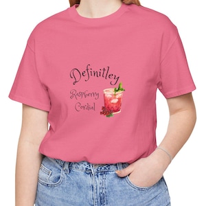 Può includere: Una t-shirt rosa a maniche corte con una grafica di un bicchiere di cordiale al lampone e la scritta "Definitley Raspberry Cordial" stampata in nero. La maglietta è abbinata a un paio di jeans blu, creando un outfit semplice e casual.