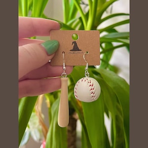 May include: Baseball-themed earrings. One earring features a miniature wooden baseball bat, and the other a white baseball with red stitching. The earrings are displayed on a brown tag with the text "SPELLBOUNDCLO".