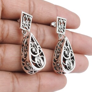 May include: A pair of silver filigree drop earrings. Each earring features a rectangular top with an openwork design, connected to a teardrop-shaped pendant with intricate scrollwork. The earrings are displayed against a light background.