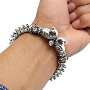 May include: Silver bangle bracelet with intricate detailing. The bracelet has small red gemstones and decorative elements. The clasp is designed with two bird-like figures. The bracelet is worn on a wrist.