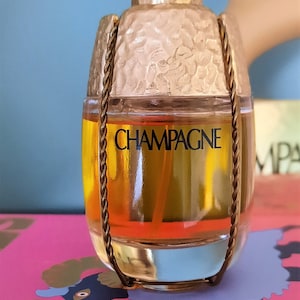 Könnte beinhalten: Eine goldene Flasche YSL Champagne Parfum mit einer strukturierten Glasflasche und einem goldenen Kettendetail. Die Flasche steht auf einer rosa Oberfläche mit einem schwarz-weißen Design.