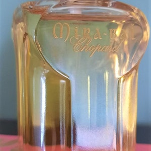 Könnte beinhalten: Eine durchsichtige Glasflasche mit Mira-B by Chopard Parfum, goldener Kappe und goldenem Lotusblüten-Logo auf dem Boden.