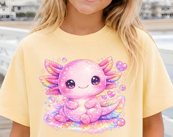 May include: A pale yellow t-shirt featuring a graphic print of a cute, pink, cartoon axolotl surrounded by colorful bubbles and small hearts. The shirt has short sleeves and a crew neckline. The text "BUTTER" is visible in a small box.