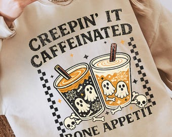 May include: A sand-colored crewneck sweatshirt featuring a graphic print of two coffee cups with ghost illustrations, spiderwebs, and skulls. The text reads "CREEPIN' IT CAFFEINATED" and "BONE APPETIT" in black lettering, framed by a black and white checkered border.