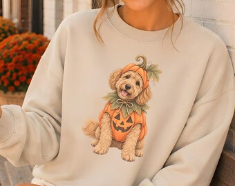 May include: A beige crewneck sweatshirt featuring a graphic illustration of a light brown puppy wearing a pumpkin costume. The puppy has a green stem hat and a carved jack-o'-lantern body suit. The sweatshirt has long sleeves and a ribbed collar.