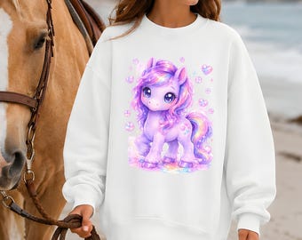 May include: A white long-sleeve sweatshirt featuring a graphic print of a purple pony with a rainbow mane and tail, surrounded by small pink hearts and bubbles. The sweatshirt is worn with a beige cowboy hat and brown leather cowboy boots.