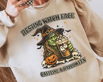May include: A beige crewneck sweatshirt featuring a graphic of a green-skinned witch wearing a hat, holding a coffee cup with ghost designs, surrounded by a pumpkin, skulls, and bones, with the text Resting Witch Face and Caffeine & Halloween.