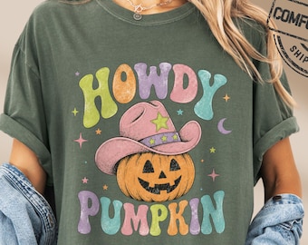 May include: A moss green short-sleeve t-shirt featuring a graphic of a jack-o'-lantern wearing a pink cowboy hat. The text HOWDY PUMPKIN is printed in colorful, wavy letters above and below the pumpkin. Small stars and a crescent moon surround the design.