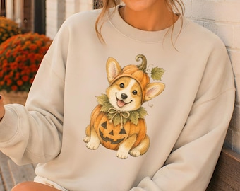 May include: A beige crewneck sweatshirt featuring a graphic illustration of a Corgi puppy wearing a pumpkin costume with a green stem and leaves. The puppy has a smiling expression and is positioned in the center of the garment. The sweatshirt has long sleeves and a ribbed neckline, cuffs, and hem.