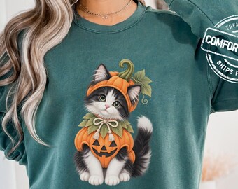 May include: A dark green long-sleeve sweatshirt featuring a graphic of a black and white kitten wearing a pumpkin costume with a jack-o'-lantern face and green leaves. The upper right sleeve includes a circular logo with the text COMFORT COLORS.