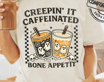 May include: An ivory t-shirt featuring a graphic print with the text "CREEPIN' IT CAFFEINATED" and "BONE APPETIT". The design includes two cups of coffee, ghosts, spiderwebs, and skulls, framed by a black and white checkered border pattern.