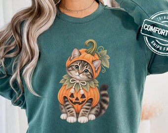 May include: A dark green long-sleeve sweatshirt featuring a graphic of a tabby cat wearing a pumpkin costume with a jack-o'-lantern face. The text COMFORT COLORS is printed in a circular badge on the upper right sleeve. The fabric has a vintage wash.