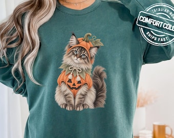 May include: A dark green long-sleeve sweatshirt featuring a graphic of a fluffy cat wearing a pumpkin costume with a jack-o'-lantern face. The upper right corner includes a circular logo with the text COMFORT COLORS.
