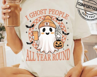 May include: Ivory-colored t-shirt with a Halloween-themed graphic. The design features a cartoon ghost wearing a beanie and holding a coffee cup and a jack-o'-lantern. The text reads "I GHOST PEOPLE ALL YEAR ROUND".