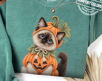 May include: A sage green crewneck sweatshirt featuring a graphic of a Siamese kitten wearing a pumpkin costume with a jack-o'-lantern face on the chest. The kitten has blue eyes and is surrounded by green leaves. A Comfort Colors label is visible.