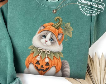 May include: A sage green crewneck sweatshirt featuring a graphic of a fluffy cat wearing a pumpkin costume with a jack-o'-lantern face and green leaves. The garment has a Comfort Colors brand label at the inner neckline and a circular logo stamp.