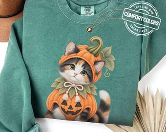 May include: A sage green crewneck sweatshirt featuring a graphic of a calico kitten wearing a pumpkin costume. The kitten has a pumpkin hat and a pumpkin body suit with a jack-o'-lantern face. The sweatshirt has a Comfort Colors brand label.
