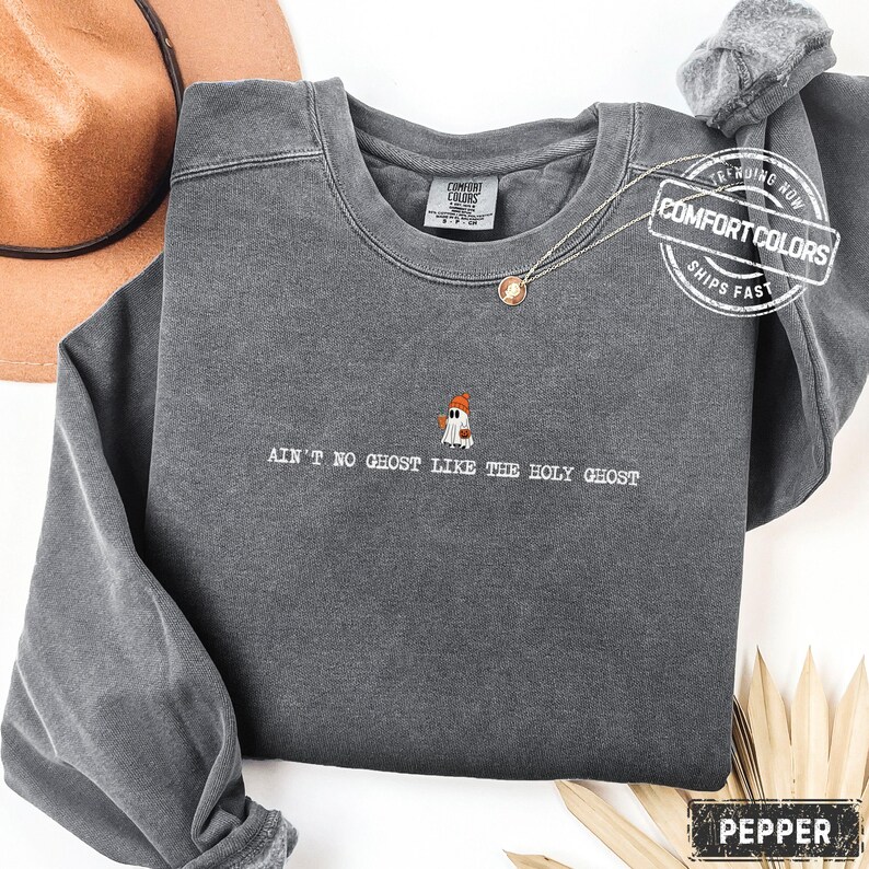 May include: A grey Comfort Colors sweatshirt featuring the text "AIN'T NO GHOST LIKE THE HOLY GHOST" and a small ghost graphic. The sweatshirt has a crew neck and long sleeves. A gold necklace with a pendant is visible.