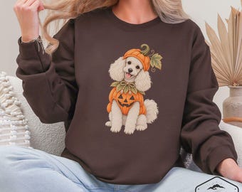 May include: A dark brown crewneck sweatshirt featuring a graphic of a white poodle wearing a pumpkin hat and a pumpkin-shaped costume with a jack-o'-lantern face. The dog has a green leaf accent on its head and a tied ribbon collar around its neck.
