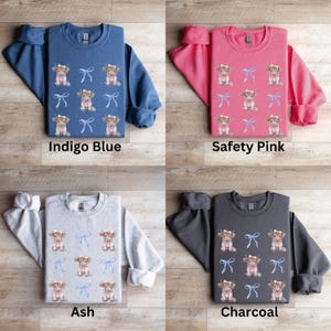May include: Four crewneck sweatshirts with a Highland cow pattern. The sweatshirts are in indigo blue, safety pink, ash grey, and charcoal. Each sweatshirt features a repeating pattern of a Highland cow with a blue bow.
