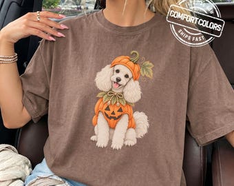 May include: A brown short-sleeved t-shirt featuring a graphic of a white poodle wearing a pumpkin hat and a pumpkin-shaped outfit. The shirt includes a circular logo in the top right corner with the text COMFORT COLORS and the word ESPRESSO at the bottom.