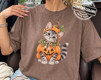 May include: A brown short-sleeve t-shirt featuring a graphic of a gray tabby cat wearing a pumpkin costume with a jack-o'-lantern face. The cat has a green collar with a bow. The shirt includes a circular logo that reads COMFORT COLORS.
