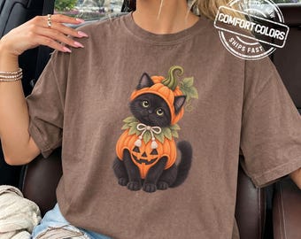 May include: A brown short-sleeve t-shirt featuring a graphic of a black cat wearing a pumpkin costume with a green stem and leaves. The cat has a collar with a bow and tassels. The shirt has a round neckline and a relaxed fit.