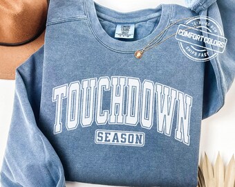 May include: A blue jean-coloured sweatshirt with the words "TOUCHDOWN SEASON" in white block letters. The sweatshirt has a crew neck and long sleeves. A gold necklace with a pendant is draped on the sweatshirt. A brown hat is in the background.