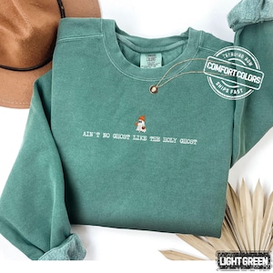 May include: A light green Comfort Colors sweatshirt with the phrase "AIN'T NO GHOST LIKE THE HOLY GHOST" embroidered below a ghost wearing a hat. The sweatshirt has a crew neck and long sleeves. A gold necklace is draped on the front.