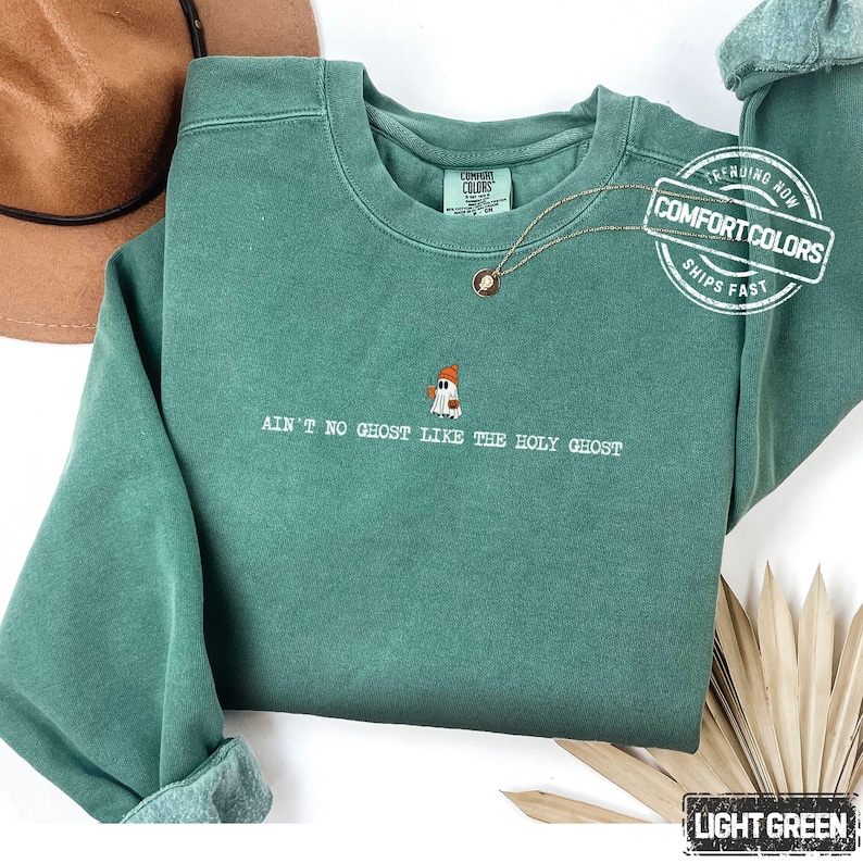 May include: A light green Comfort Colors sweatshirt with the phrase "AIN'T NO GHOST LIKE THE HOLY GHOST" embroidered below a ghost wearing a hat. The sweatshirt has a crew neck and long sleeves. A gold necklace is draped on the front.