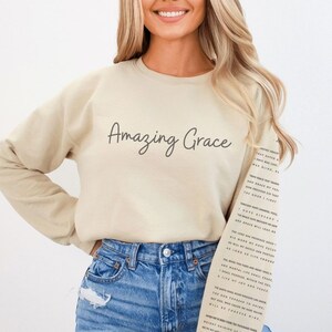 Op de afbeelding: Een beige sweatshirt met de tekst "Amazing Grace" erop gedrukt. De tekst staat in een cursief lettertype. Het sweatshirt wordt gedragen door iemand met blond haar en een blauwe jeans. De tekst van het kerklied "Amazing Grace" is op het sweatshirt gedrukt.