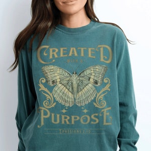 May include: Teal long-sleeved shirt with a vintage-style graphic of a butterfly and the text "CREATED WITH A PURPOSE - EPHESIANS 2:10 -"