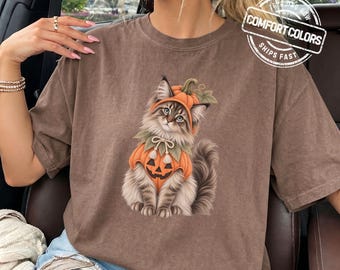 May include: A brown short-sleeved t-shirt featuring a graphic of a fluffy cat wearing an orange pumpkin costume with a jack-o'-lantern face. The cat has long fur and is sitting upright. The shirt has a crew neckline and a relaxed fit.