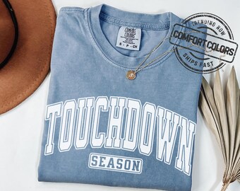 May include: A blue Comfort Colors t-shirt with the words "TOUCHDOWN SEASON" in white block letters. A gold necklace with a pendant rests on the shirt. The shirt is folded on a white surface.