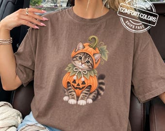 May include: A brown short-sleeve t-shirt featuring a graphic of a tabby kitten wearing a pumpkin costume with a jack-o'-lantern face, green leaves, and a bow. The shirt includes a circular logo in the top right corner with the text COMFORT COLORS.